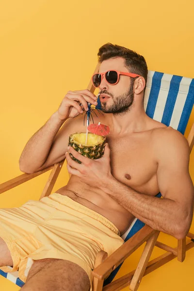 Shirtless man in sunglasses drinking fresh cocktail from half of pineapple while relaxing in deck chair isolated on yellow — Stock Photo