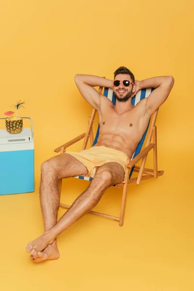 Homem sem camisa sorridente em óculos de sol descansando em cadeira de praia perto de geladeira portátil e metade de abacaxi com coquetel fresco no fundo amarelo — Fotografia de Stock