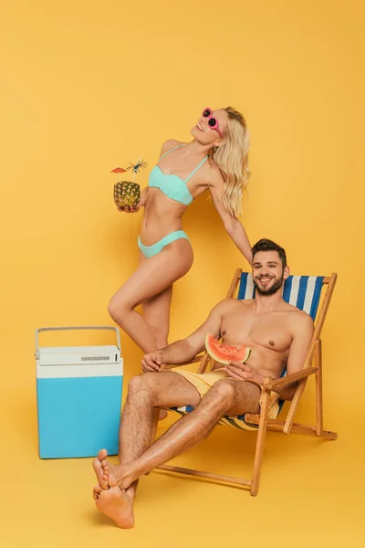 Jolie fille blonde tenant l'ananas avec cocktail près d'un réfrigérateur portable, et un homme souriant assis sur une chaise longue sur fond jaune — Photo de stock