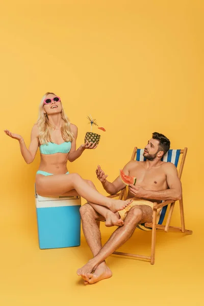 Aufgeregtes blondes Mädchen sitzt auf tragbarem Kühlschrank neben gutaussehendem Mann mit Scheibe Wassermelone im Liegestuhl auf gelbem Hintergrund — Stockfoto