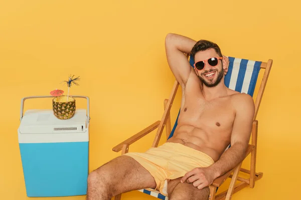 Hombre feliz sin camisa en gafas de sol descansando en tumbonas cerca de nevera portátil y la mitad de la piña con cóctel fresco sobre fondo amarillo - foto de stock