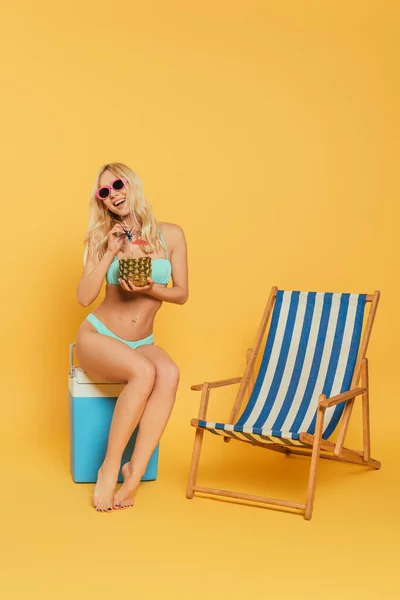 Cheerful, blonde girl with fresh cocktail in pineapple sitting on portable fridge near deck chair on yellow background — Stock Photo
