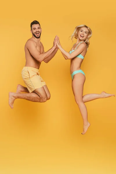Feliz joven pareja levitando con las manos unidas mientras sonríe a la cámara sobre fondo amarillo - foto de stock