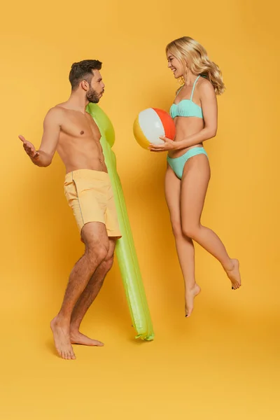 Alegre chica saltando con inflable bola cerca excitado hombre celebración inflable colchón en amarillo fondo - foto de stock