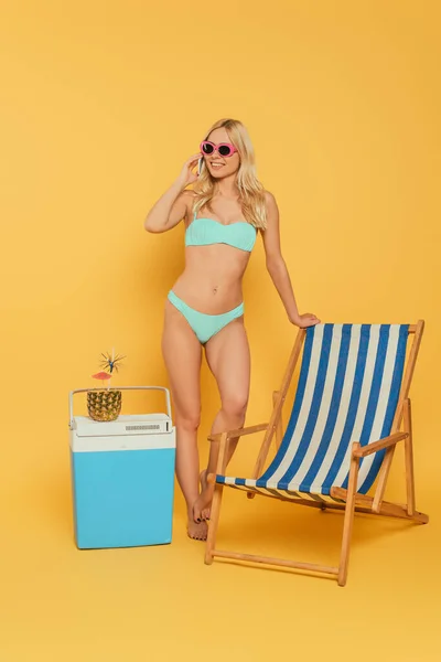 Smiling, blonde girl talking on smartphone near deck chair and portable fridge with fresh cocktail in pineapple on yellow background — Stock Photo