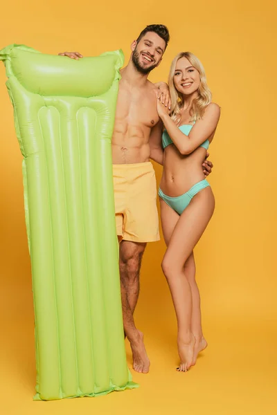 Heureux musclé homme tenant matelas gonflable et câlin attrayant fille blonde sur fond jaune — Photo de stock