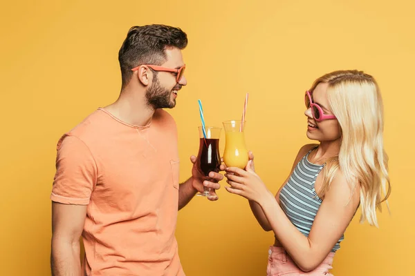 Sourire jeune couple dans des lunettes de soleil cliquetis verres à cocktail sur fond jaune — Photo de stock