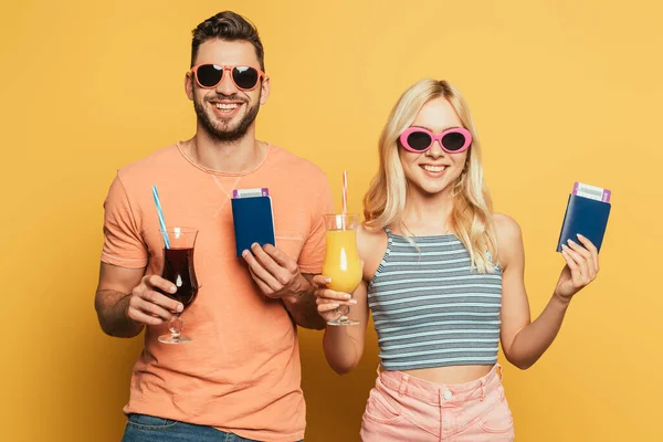 Allegra giovane coppia in occhiali da sole che tiene cocktail e documenti mentre sorride alla fotocamera su sfondo giallo — Foto stock