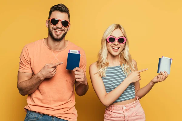 Feliz pareja joven en gafas de sol apuntando con los dedos a los pasaportes y billetes de avión sobre fondo amarillo - foto de stock