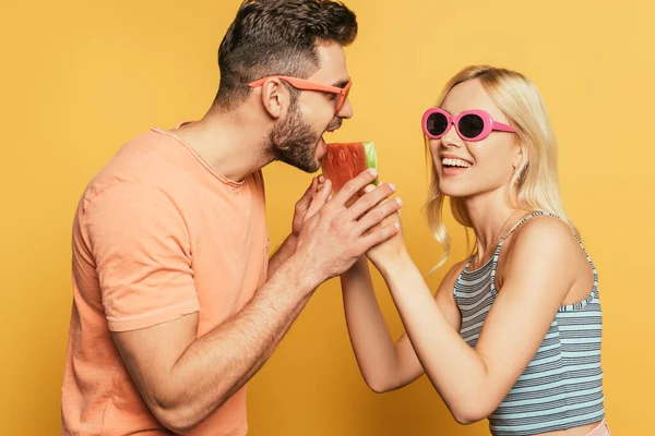 Cheerful blonde girl feeding handsome boyfriend with juicy watermelon on yellow background — Stock Photo