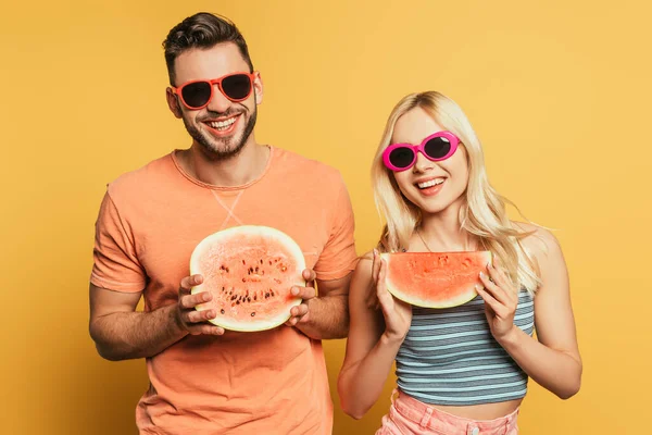 Happy young couple in sunglasses showing cut, ripe watermelon on yellow background — Stock Photo