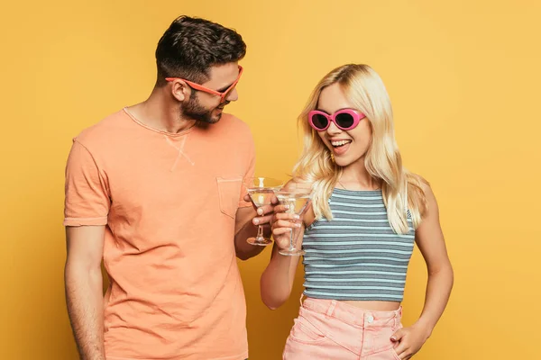 Glückliches junges Paar in Sonnenbrille klimpert Cocktailgläser auf gelbem Hintergrund — Stockfoto