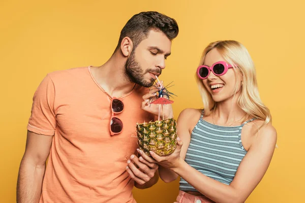 Schöner Mann trinkt Cocktail aus Ananas in den Händen eines lächelnden blonden Mädchens auf gelbem Hintergrund — Stockfoto