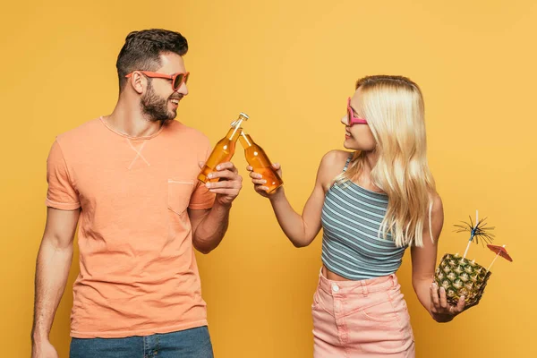 Bionda ragazza tenendo cocktail in ananas mentre clinking bottiglie di birra con sorridente fidanzato su sfondo giallo — Foto stock