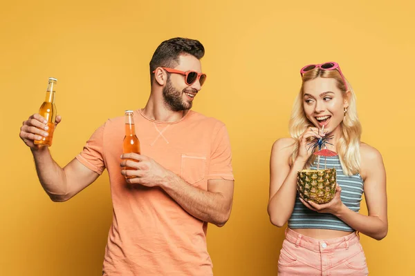 Uomo sorridente in possesso di bottiglie di birra vicino allegra ragazza bere cocktail da ananas su sfondo giallo — Foto stock