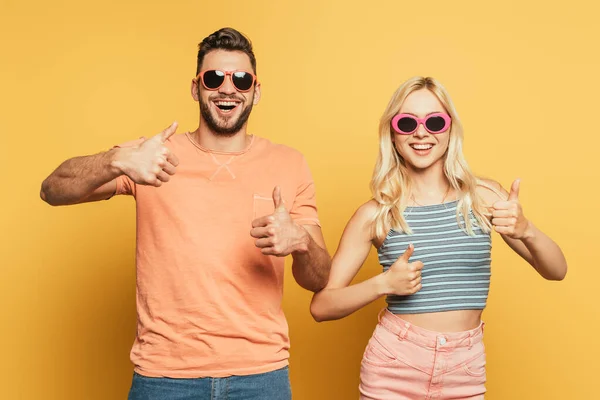 Весела пара в сонцезахисних окулярах показує великі пальці вгору, дивлячись на камеру на жовтому фоні — стокове фото