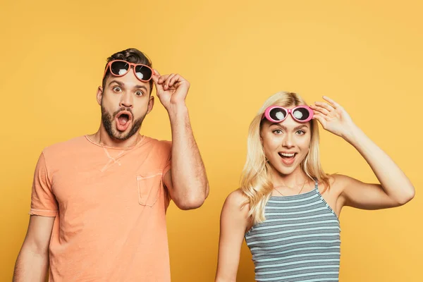 Conmocionados hombre y mujer tocando gafas de sol mientras mira a la cámara en el fondo amarillo - foto de stock