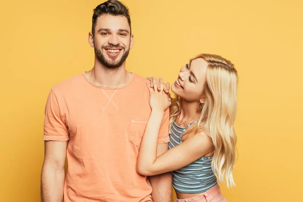 Glückliches blondes Mädchen umarmt Schulter eines lächelnden Mannes isoliert auf gelb — Stockfoto