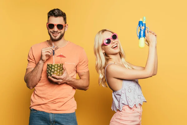 Menina loira alegre segurando arma de água perto de homem sorridente bebendo coquetel de metade de abacaxi no fundo amarelo — Fotografia de Stock