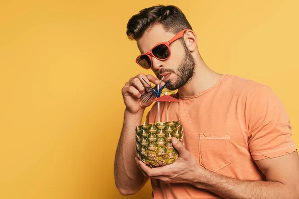 Joven confiado en gafas de sol bebiendo cóctel fresco de la mitad de la piña fresca sobre fondo amarillo - foto de stock