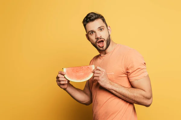 Sorprendido joven sosteniendo rebanada de jugosa sandía mientras mira a la cámara en el fondo amarillo - foto de stock