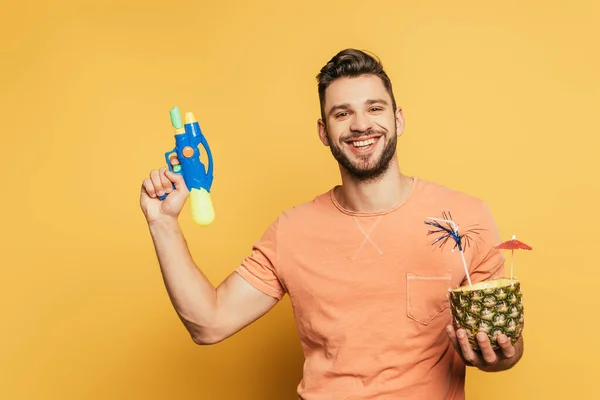 Homme joyeux tenant pistolet à eau et la moitié de l'ananas avec cocktail frais sur fond jaune — Photo de stock