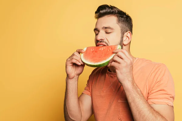 Schöner junger Mann mit geschlossenen Augen isst reife, saftige Wassermelone auf gelbem Hintergrund — Stockfoto