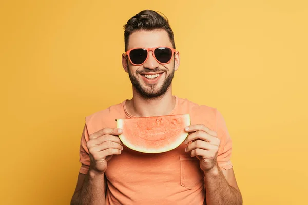 Happy young man in sunglasses holding slice of ripe watermelon on yellow background — Stock Photo