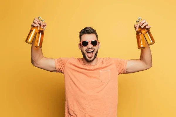 Jovem animado em óculos de sol segurando garrafas de cerveja enquanto olha para a câmera no fundo amarelo — Fotografia de Stock