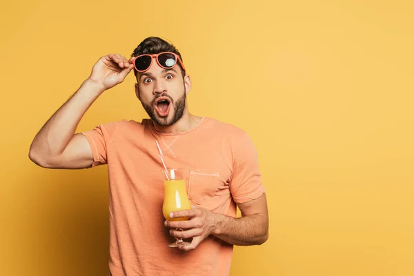 Chocado jovem com boca aberta tocando óculos de sol enquanto segurando suco de laranja no fundo amarelo — Fotografia de Stock