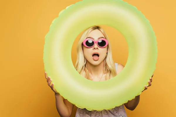 Choqué fille blonde avec bouche ouverte regardant à travers anneau de nage sur fond jaune — Photo de stock