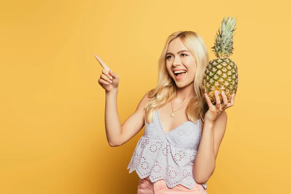 Cheerful blonde girl holding pineapple and pointing with finger on yellow background — Stock Photo