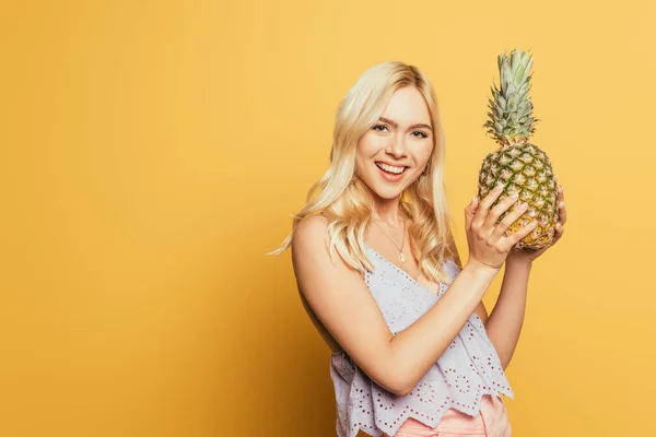 Happy blonde girl holding pineapple while smiling at camera on yellow background — Stock Photo