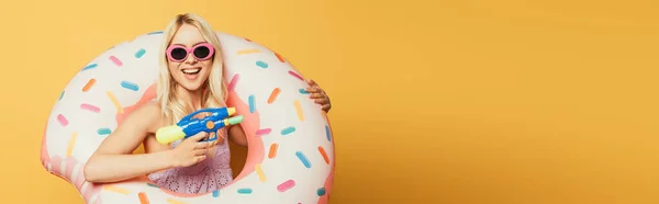 Panoramic crop of happy blonde girl in swim ring holding water gun on yellow background — Stock Photo