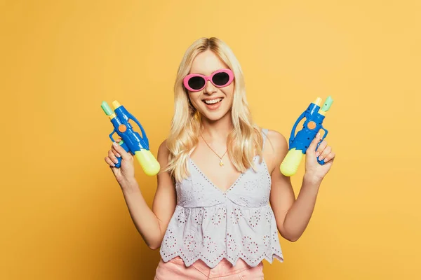 Весела блондинка в сонцезахисних окулярах тримає водяні гармати на жовтому тлі — стокове фото