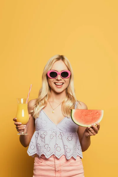 Smiling, blonde girl in sunglasses holding glass of orange juice and slice of watermelon on yellow background — Stock Photo