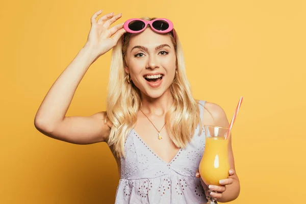 Aufgeregtes blondes Mädchen berührt Sonnenbrille, während es ein Glas Orangensaft auf gelbem Hintergrund hält — Stockfoto