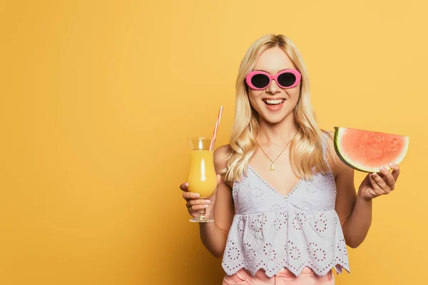 Chica rubia feliz en gafas de sol sosteniendo jugo de naranja y rebanada de sandía sobre fondo amarillo - foto de stock