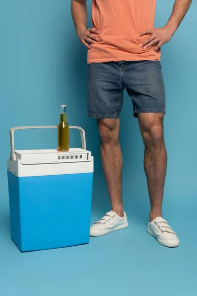 Vista ritagliata dell'uomo in piedi con le mani sui fianchi vicino bottiglia di birra sul frigorifero portatile su sfondo blu — Foto stock