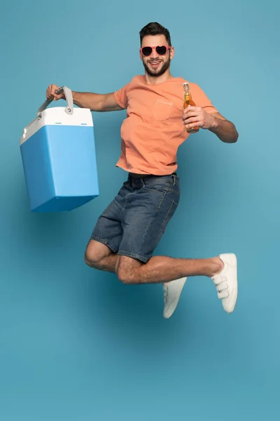 Homem animado em óculos de sol pulando enquanto segurava garrafa de cerveja e geladeira portátil no fundo azul — Fotografia de Stock