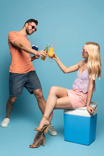 Blondes Mädchen sitzt auf tragbarem Kühlschrank und klimpert mit einem Glas Orangensaft, während der Mann eine Flasche Bier, Reisepass und Flugtickets auf blauem Hintergrund hält — Stockfoto