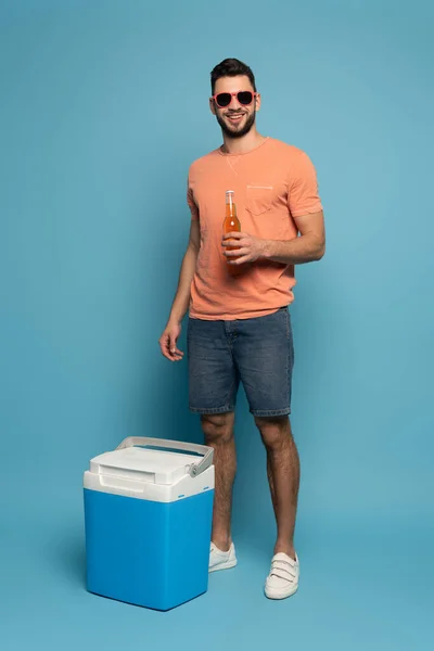 Happy man in sunglasses holding bottle of beer while standing near portable fridge on blue background — Stock Photo