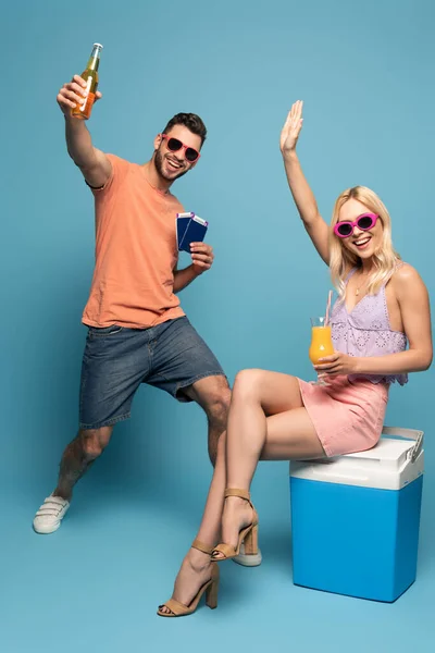 Chica feliz con un vaso de jugo de naranja sentado en la nevera portátil y agitando la mano cerca de hombre emocionado con documentos y botella de cerveza sobre fondo azul - foto de stock