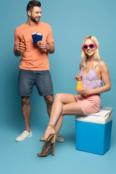 Menina feliz com vidro de suco de laranja sentado na geladeira portátil perto de homem sorridente segurando documentos e garrafa de cerveja no fundo azul — Fotografia de Stock