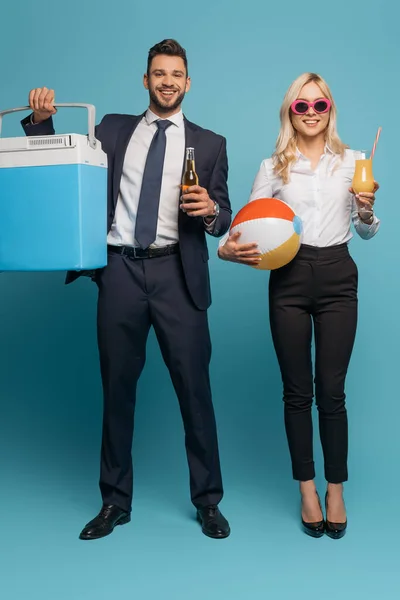 Повний вигляд на бізнесмена з портативним холодильником і пивом біля усміхненої бізнес-леді з надувним м'ячем і апельсиновим соком на синьому фоні — стокове фото