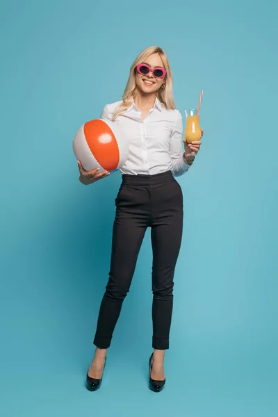 Ganzkörperansicht einer lächelnden Geschäftsfrau mit Sonnenbrille, die Orangensaft und aufblasbare Kugel auf blauem Hintergrund hält — Stockfoto