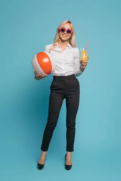 Full length view of smiling businesswoman in sunglasses holding orange juice and inflatable ball on blue background — Stock Photo