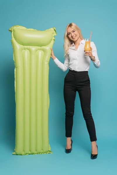 Full length view of cheerful businesswoman holding glass of orange juice near inflatable mattress on blue background — Stock Photo