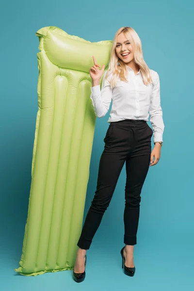 Full length view of smiling businesswoman pointing with finger at inflatable mattress on blue background — Stock Photo