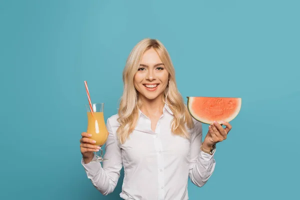 Atractiva, rubia empresaria sonriendo a la cámara blanca sosteniendo rebanada de sandía y vaso de jugo de naranja sobre fondo azul - foto de stock